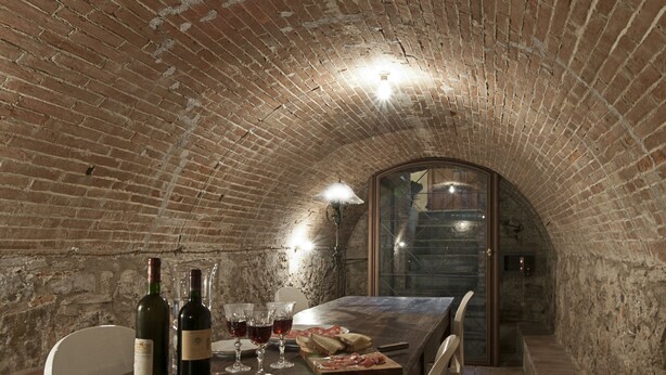 A rustic brick interior with a long table set for dining, wine bottles, glasses, and a wooden cutting board with food. - B&B VillAmorosa (Vicopisano)