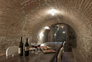 Un interior de ladrillo rústico con una larga mesa dispuesta para comer, botellas de vino, copas y una tabla de madera con comida.