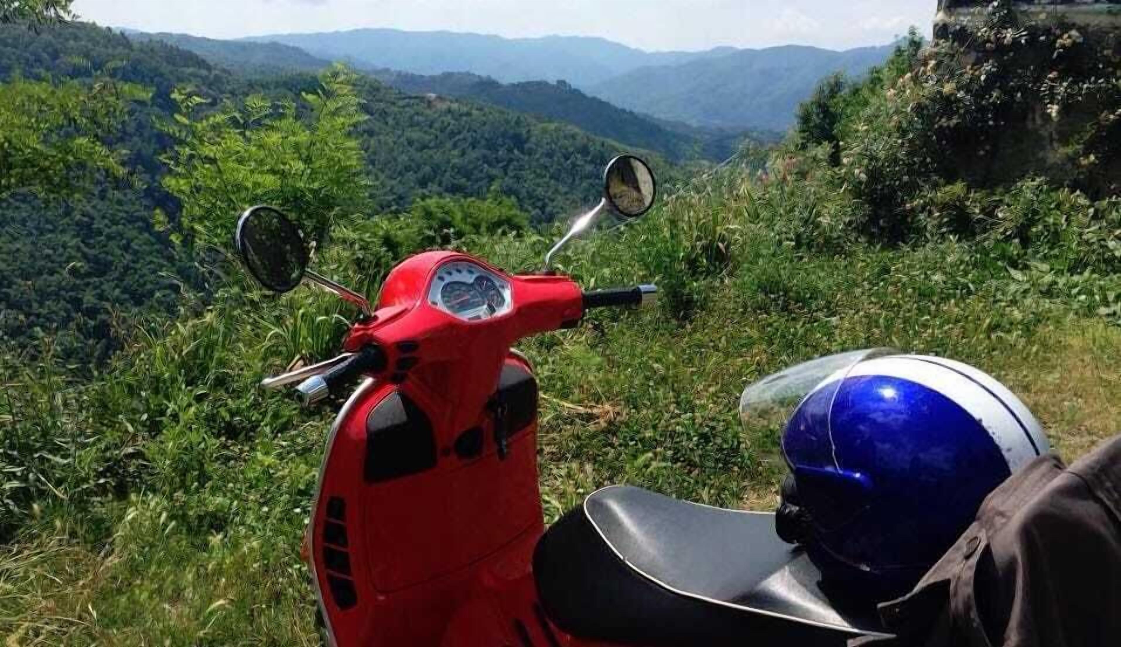 A red scooter parked on a grassy hill with a scenic view of mountains in the background.