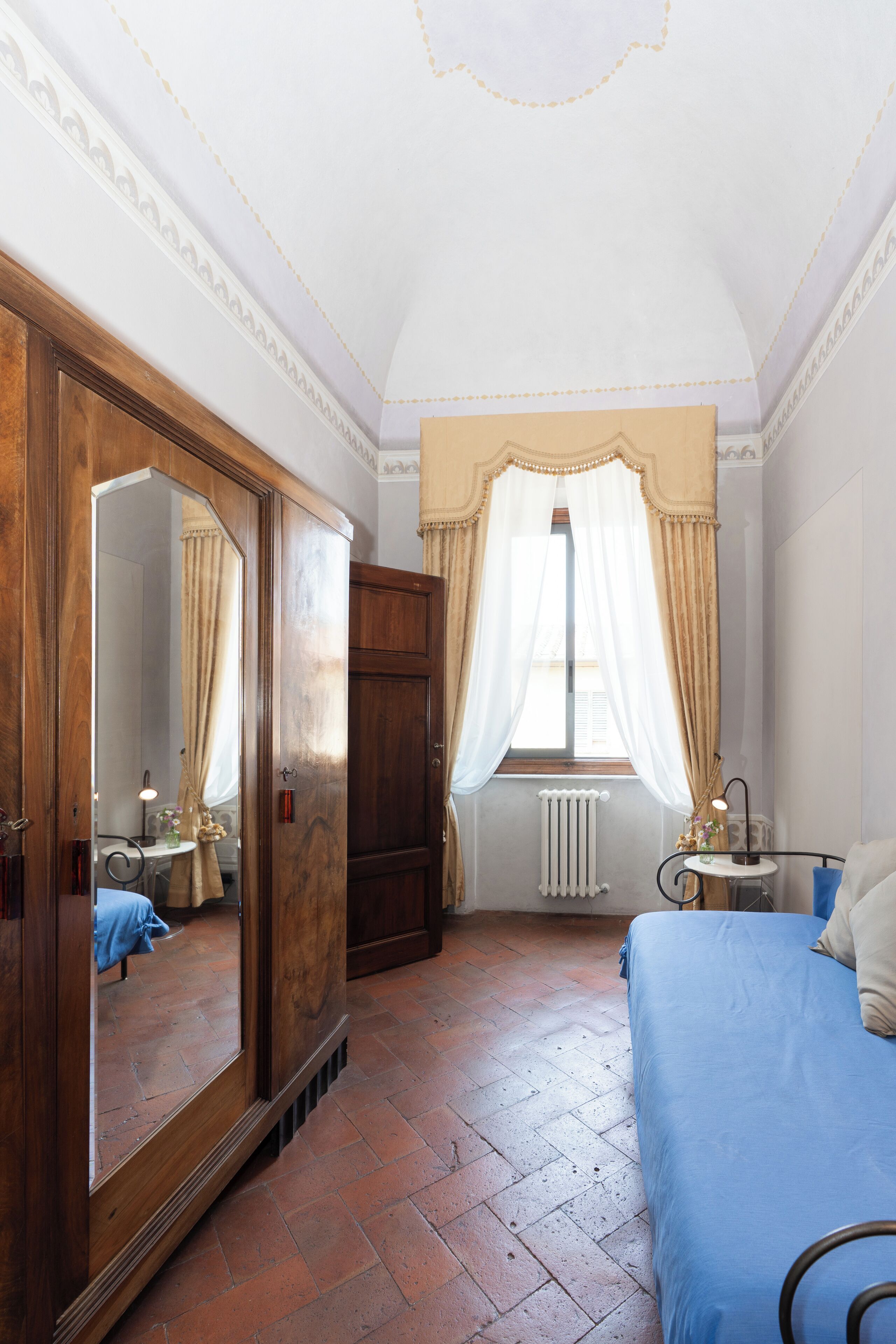 Un dormitorio con un espejo grande, un armario de madera, una cama con colcha azul y una ventana con cortinas.
