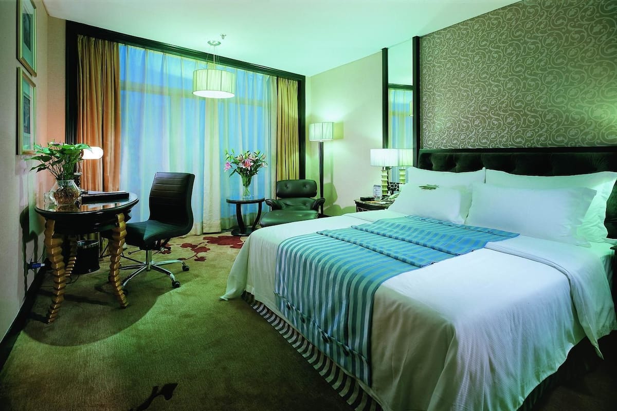 A hotel room with a large bed, a desk with a chair, a small table with a vase of flowers, and a lamp.