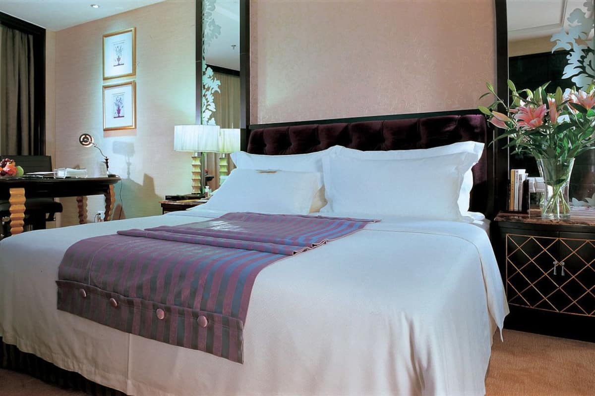 A hotel room with a large bed, a bedside table with a lamp, a small desk with a chair, and a vase with flowers on a dresser.