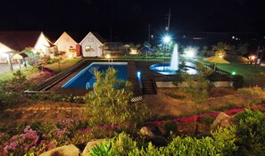 Nattebillede af et boligområde med swimmingpool, springvand og huse med oplyste vinduer.
