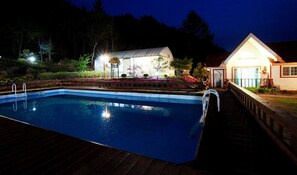En swimmingpool med træterrasse, omgivet af en have med forskellige planter og blomster. I baggrunden ses en bygning med veranda og overdækket område.