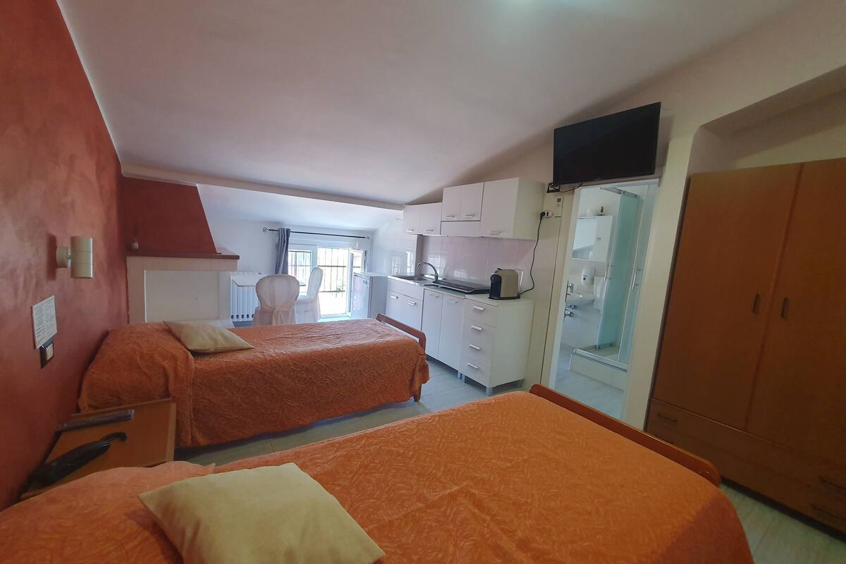 A hotel room with two beds, a kitchenette, a bathroom with a glass shower, and a TV mounted on the wall.