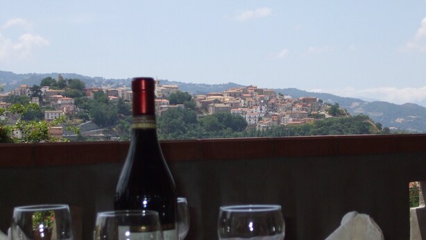A bottle of wine and three glasses on a table with a view of a town and hills in the background. - Hotel Supersonik (Acri)