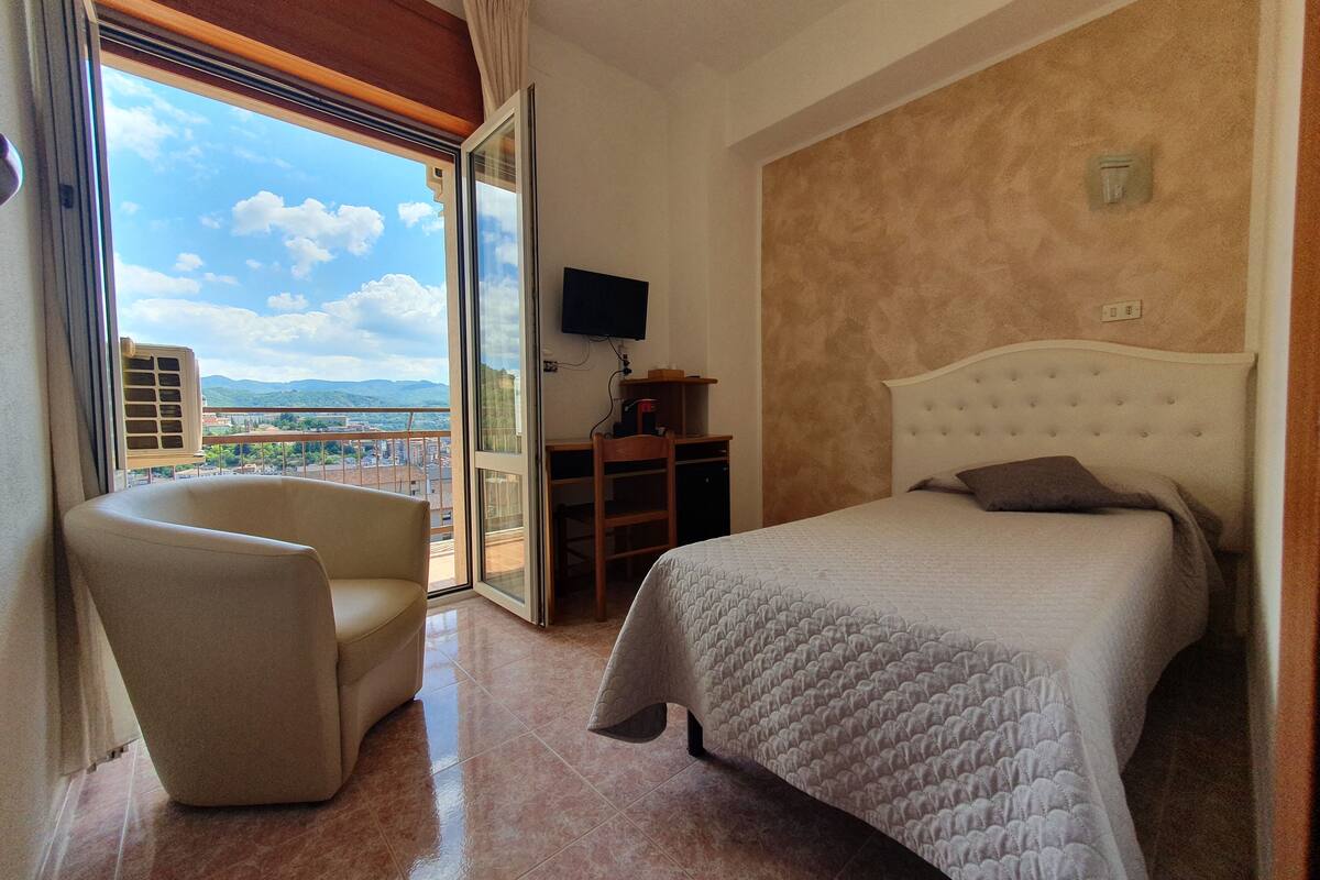 A hotel room with a bed, a chair, a desk, a TV, and a balcony with a city view.