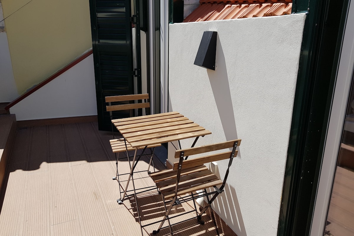 A wooden bench and table set up on a balcony with a white wall and a tiled roof in the background.