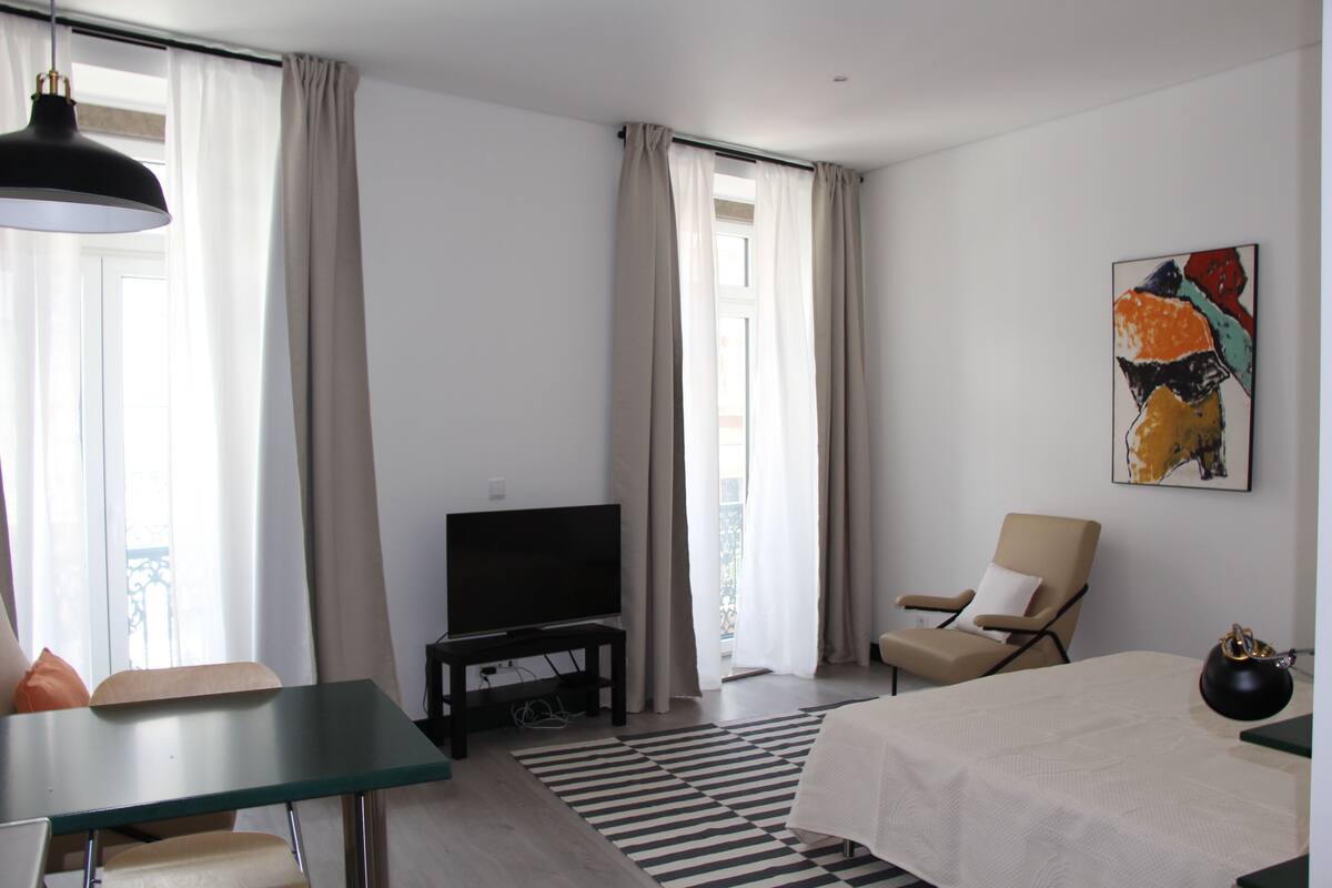 A modern hotel room with a bed, a TV on a stand, a chair, and a table.