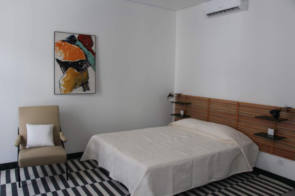 A hotel room with a bed, a chair, a wall-mounted shelf, and a checkered floor.
