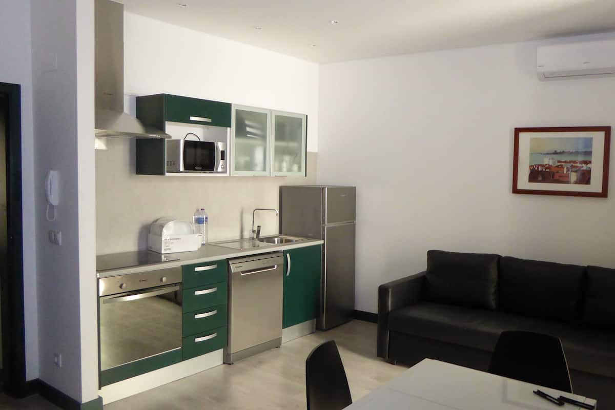 A compact kitchen with a microwave, toaster, and refrigerator. There is a dining table with chairs and a dark-colored sofa. An air conditioning unit is mounted on the wall.