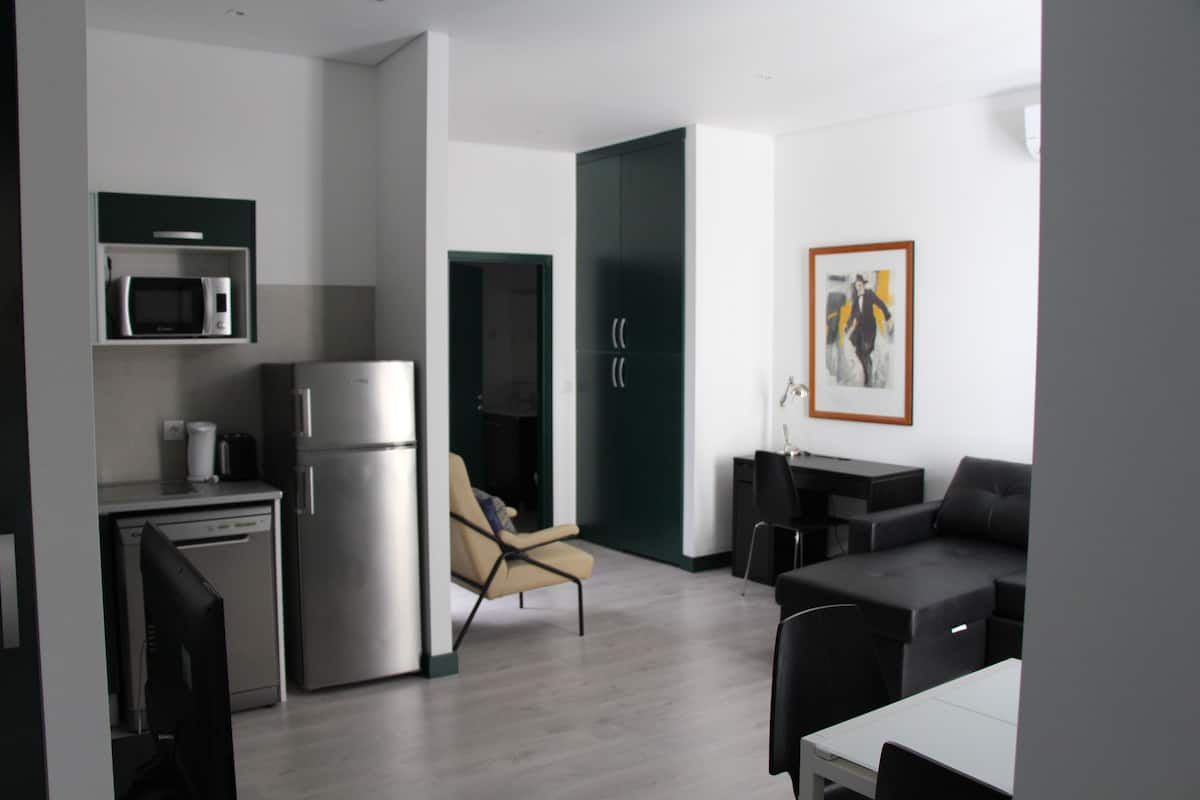 A modern kitchen with a microwave, refrigerator, and dishwasher. There is a beige chair and a black sofa in the living area.