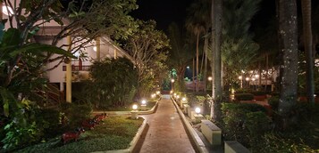 Vista nocturna de un patio de hotel con un sendero empedrado, estatuas decorativas de caballos y farolas iluminadas.