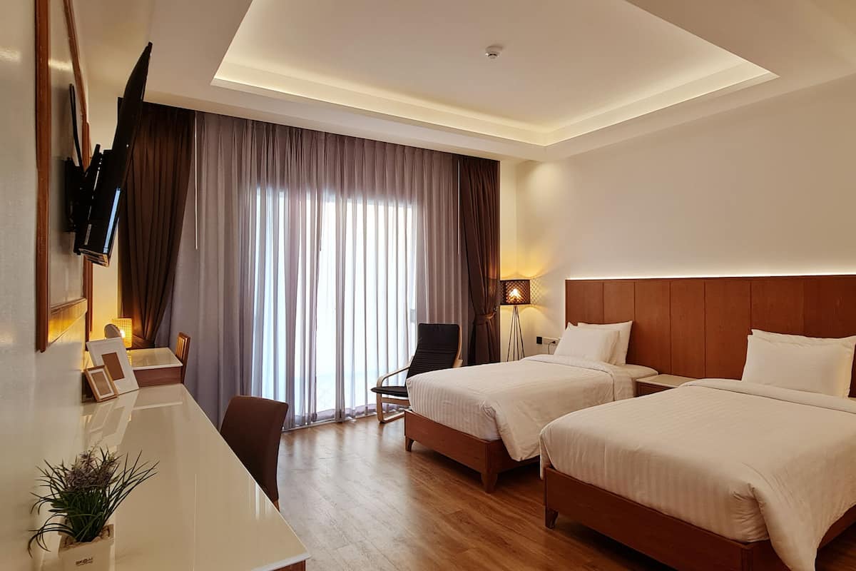 A hotel room with two beds, a desk, a chair, a TV mounted on the wall, and a window with curtains.