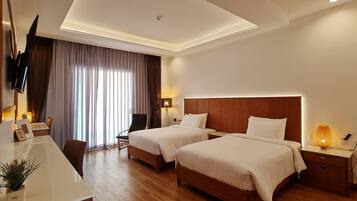 A hotel room with two beds, a desk, a chair, a TV mounted on the wall, and a window with curtains.