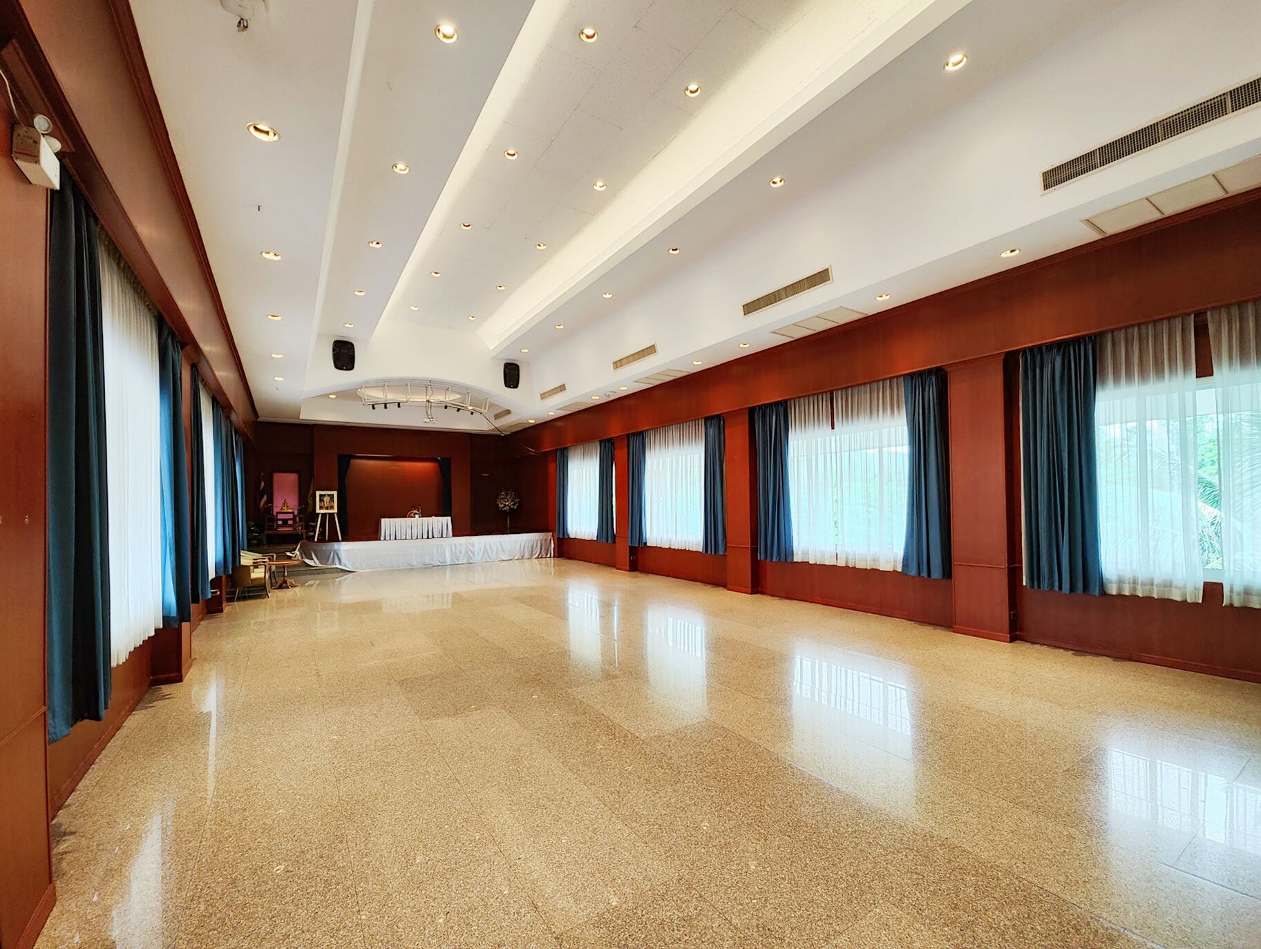 A spacious, well-lit room with a polished floor, large windows with blue curtains, and a stage area with a table and chairs.