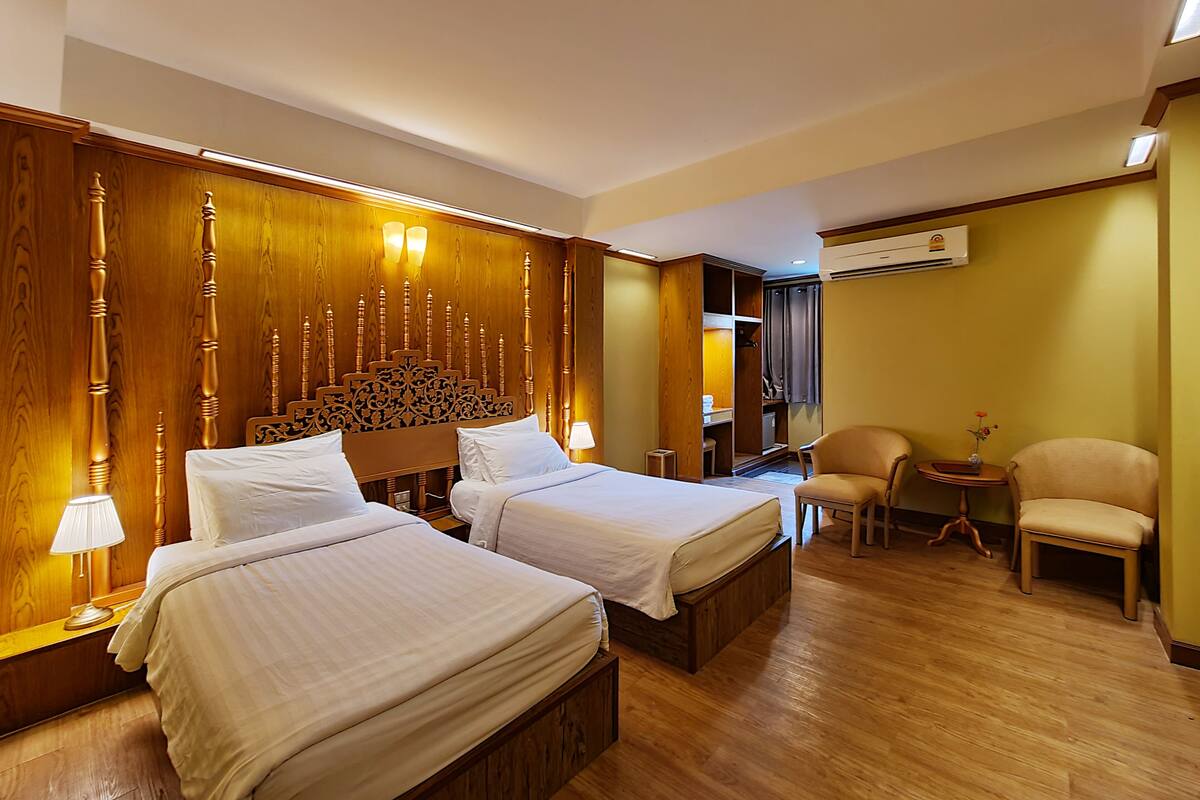 A hotel room with two beds, wooden headboard, bedside lamps, a small table, two chairs, and an air conditioning unit.