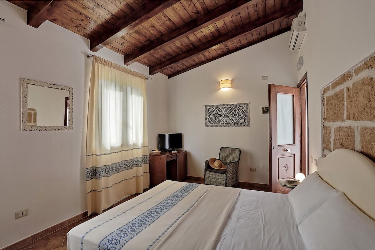 A bedroom with a wooden ceiling, a bed with a patterned bedspread, a chair, a television on a desk, and a wall-mounted light fixture.