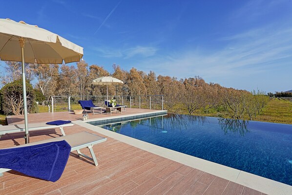 An infinity pool with lounge chairs and umbrellas, surrounded by a wooden deck and a view of trees and hills.