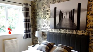 A hotel room with a bed, a window with curtains, a radiator, a bedside table with a lamp, and a wall with patterned wallpaper and a framed picture. - Abbey Grange Hotel (Nuneaton)