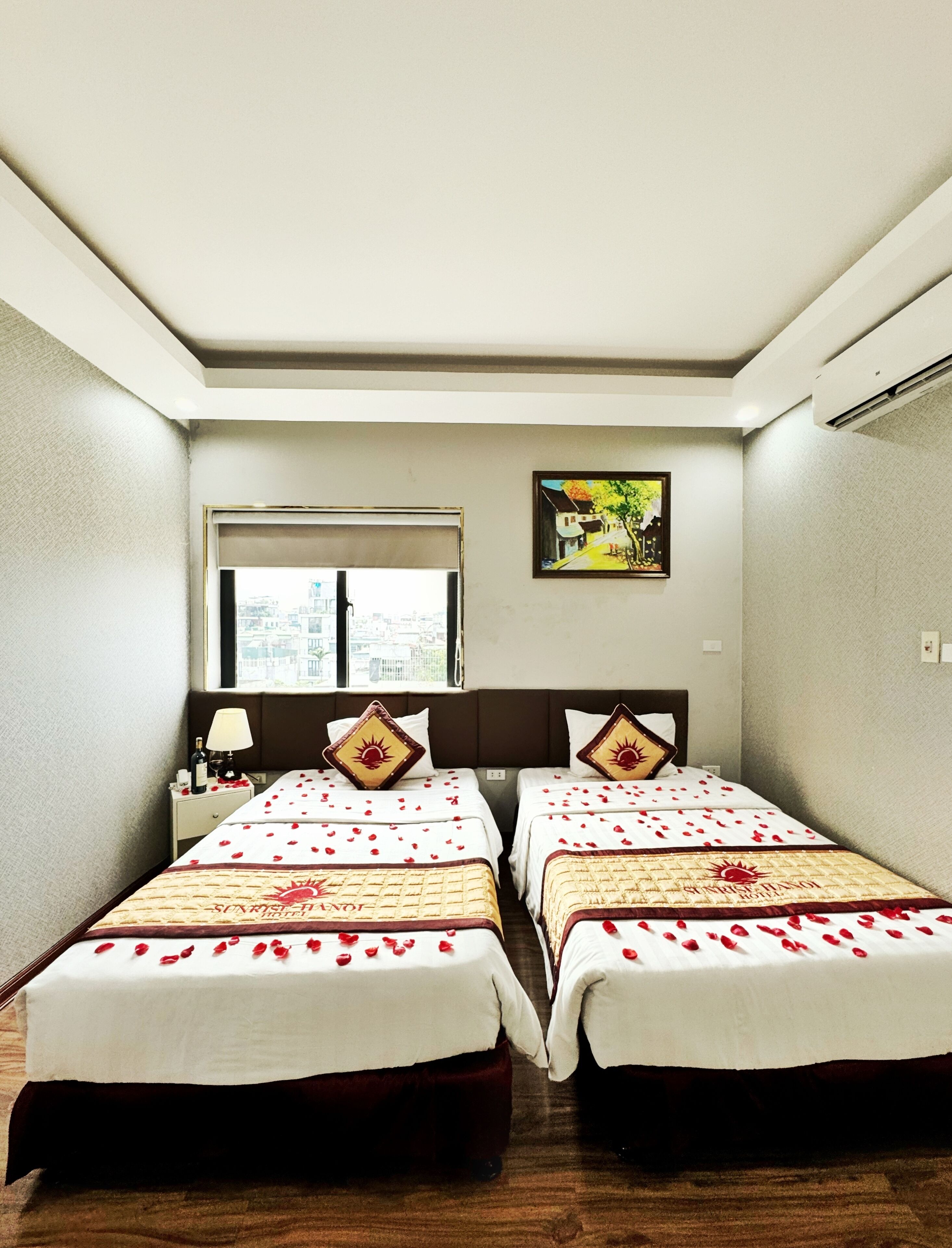 Habitación de hotel con dos camas, una ventana con vista a la ciudad, un cuadro en la pared y una mesita de noche con una lámpara y una botella.