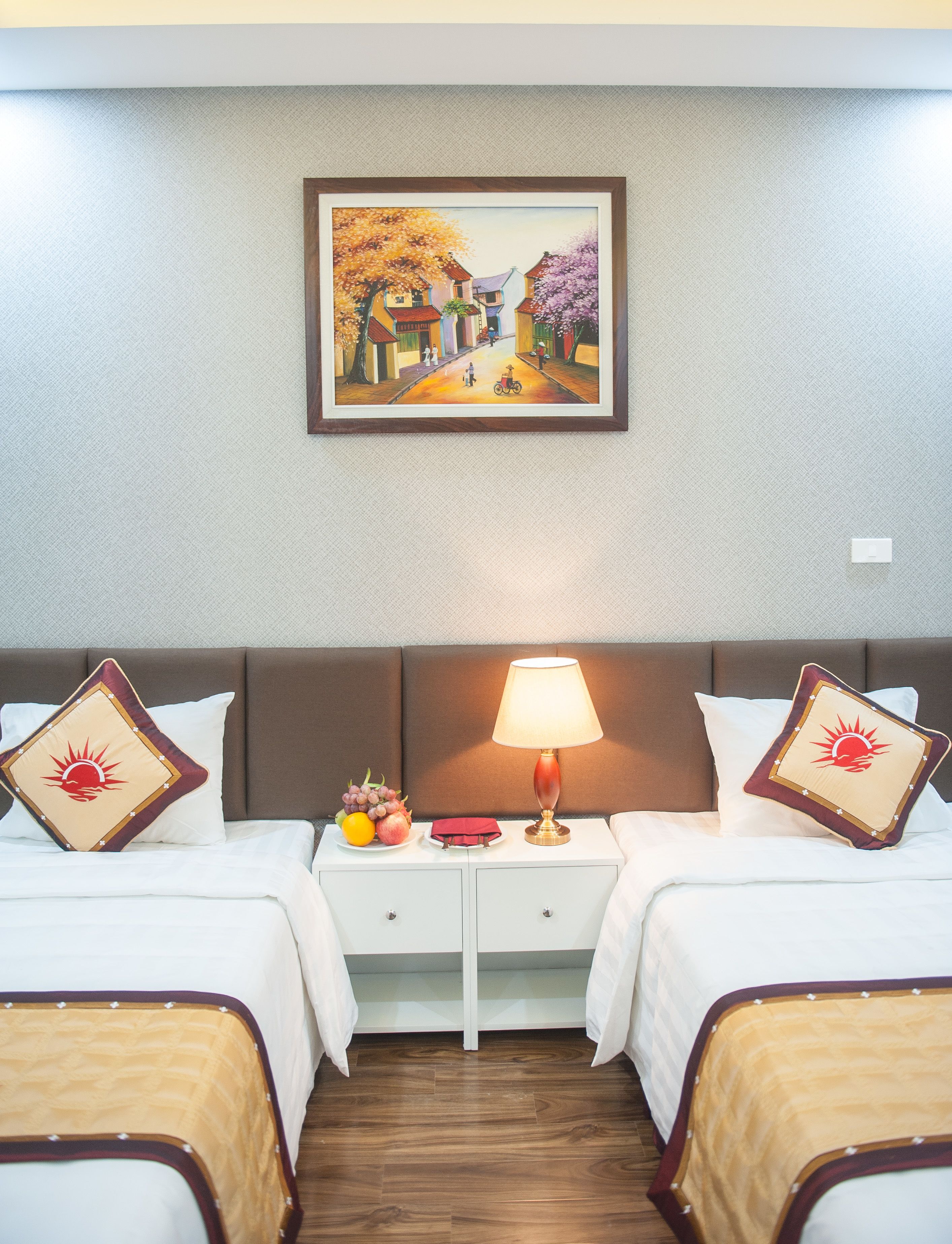 A hotel room with two beds, a nightstand with a lamp, and a painting of a village scene on the wall.