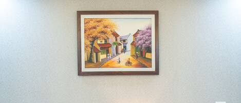 A hotel room with two beds, a nightstand with a lamp, and a painting of a village scene on the wall.