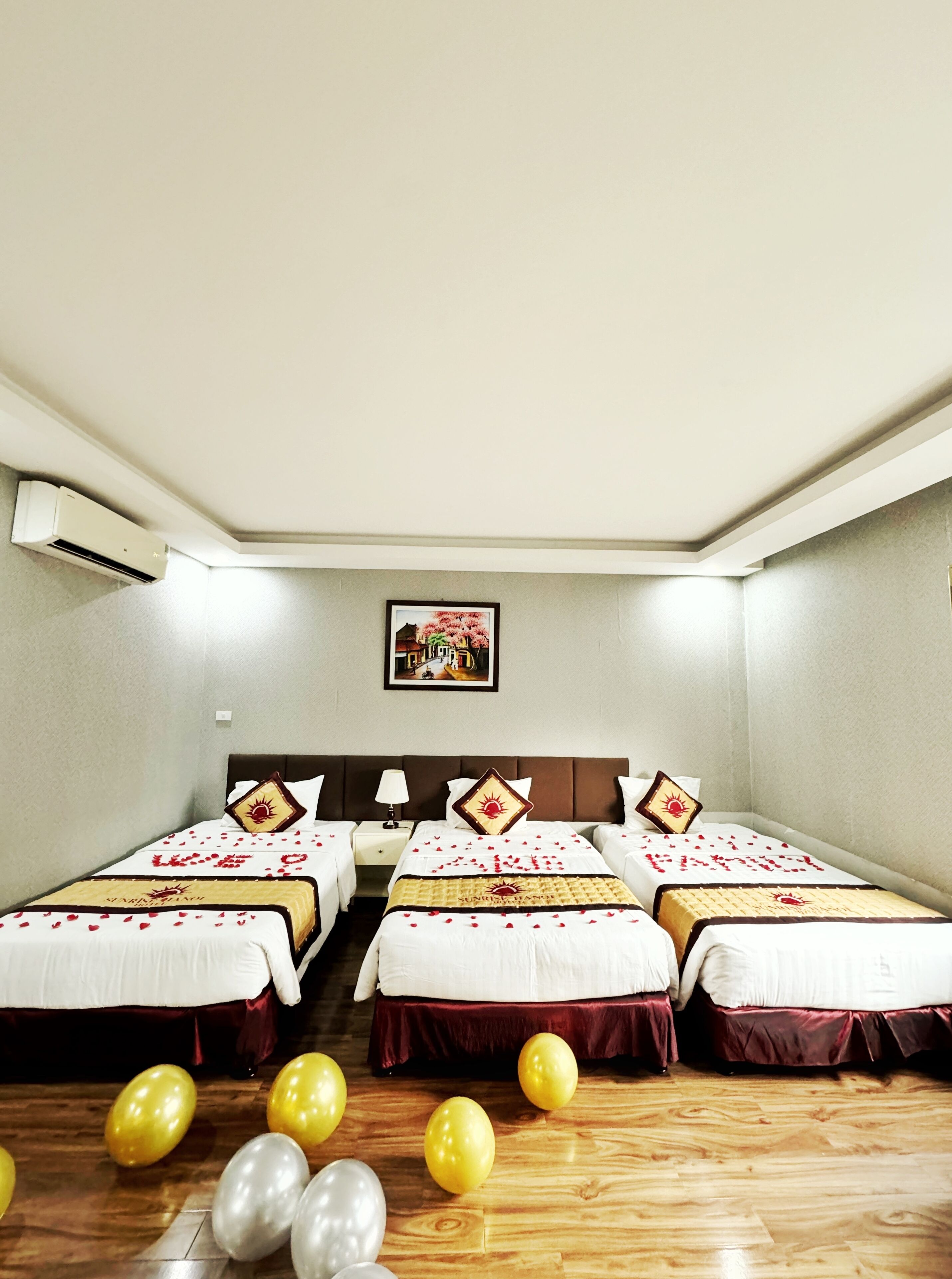 A hotel room with four beds, a ceiling fan, an air conditioner, a framed picture on the wall, and balloons on the floor.