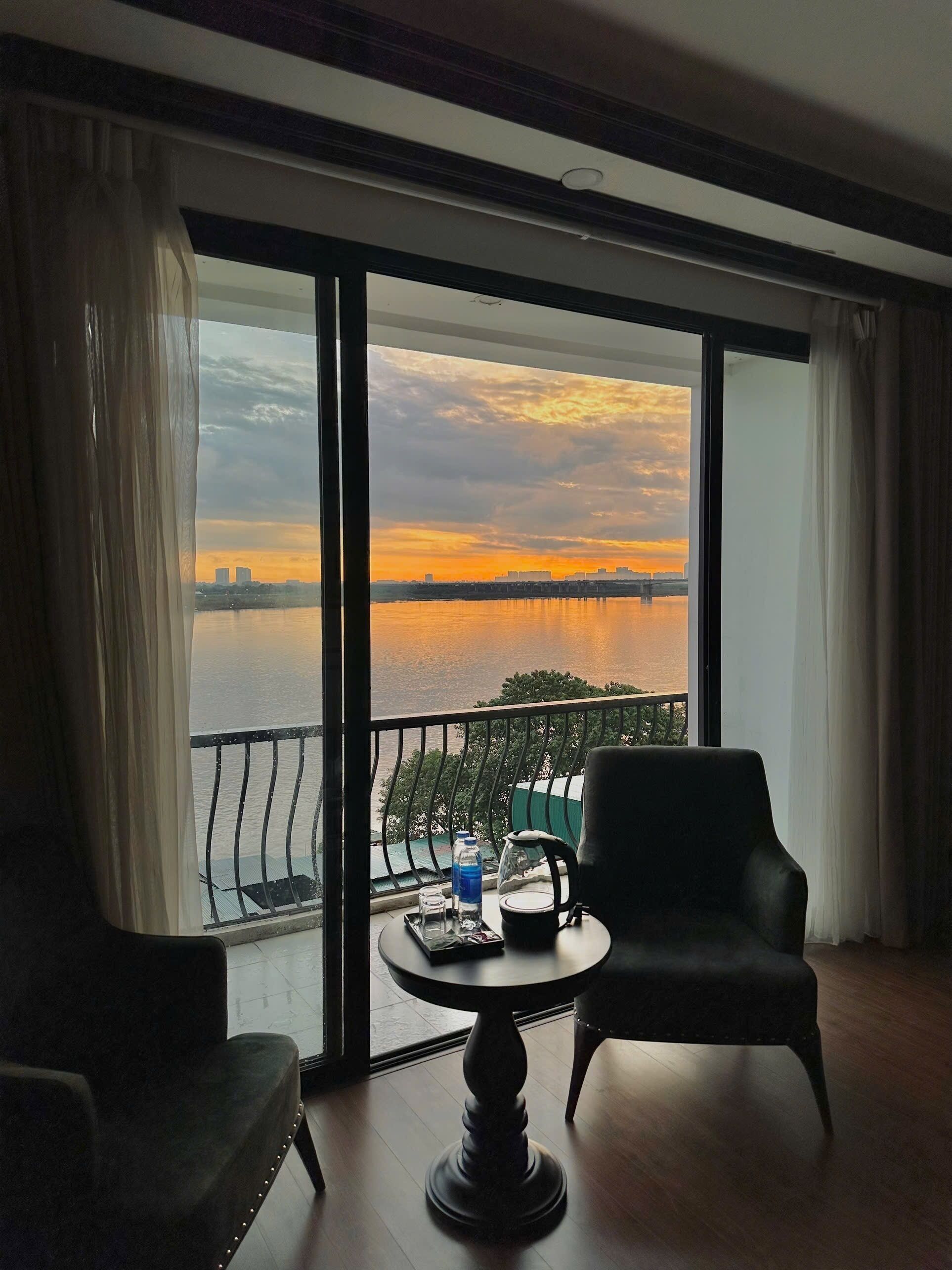 A room with a view of a sunset over a body of water, a balcony with a railing, a table with a chair, and a sofa.