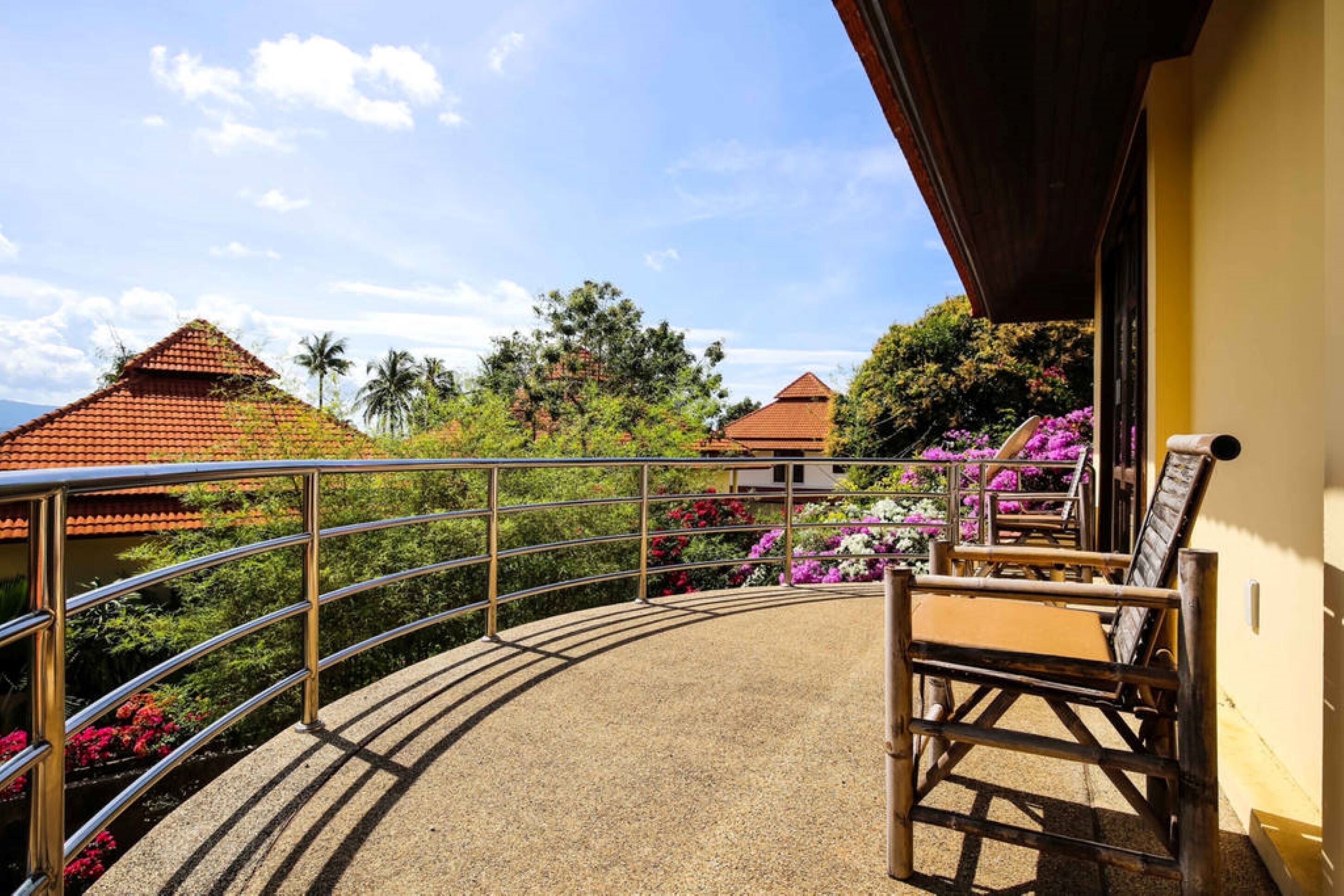 A balcony with a metal railing, a wooden chair, and a view of a building with a red roof and tropical vegetation.
