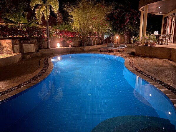 A swimming pool with a curved edge, surrounded by a stone border and a low wall. There are lounge chairs and a table with chairs on the right side.