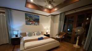 A hotel room with a large bed, bedside tables, a nightstand, a chair, a standing fan, and a painting on the wall. - 4 Bedroom Villa TG48 on beachfront resort SDV288-By Samui Dream Villas (Koh Samui)