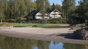Vista de un lago con playa arenosa, una mesa de picnic y un columpio. En el fondo, se ven dos casas con balcones.