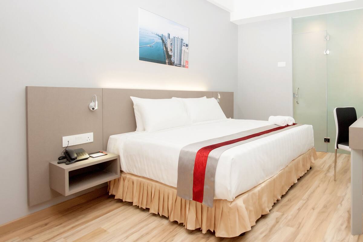 A hotel room with a bed, bedside table, desk, chair, and a painting on the wall.