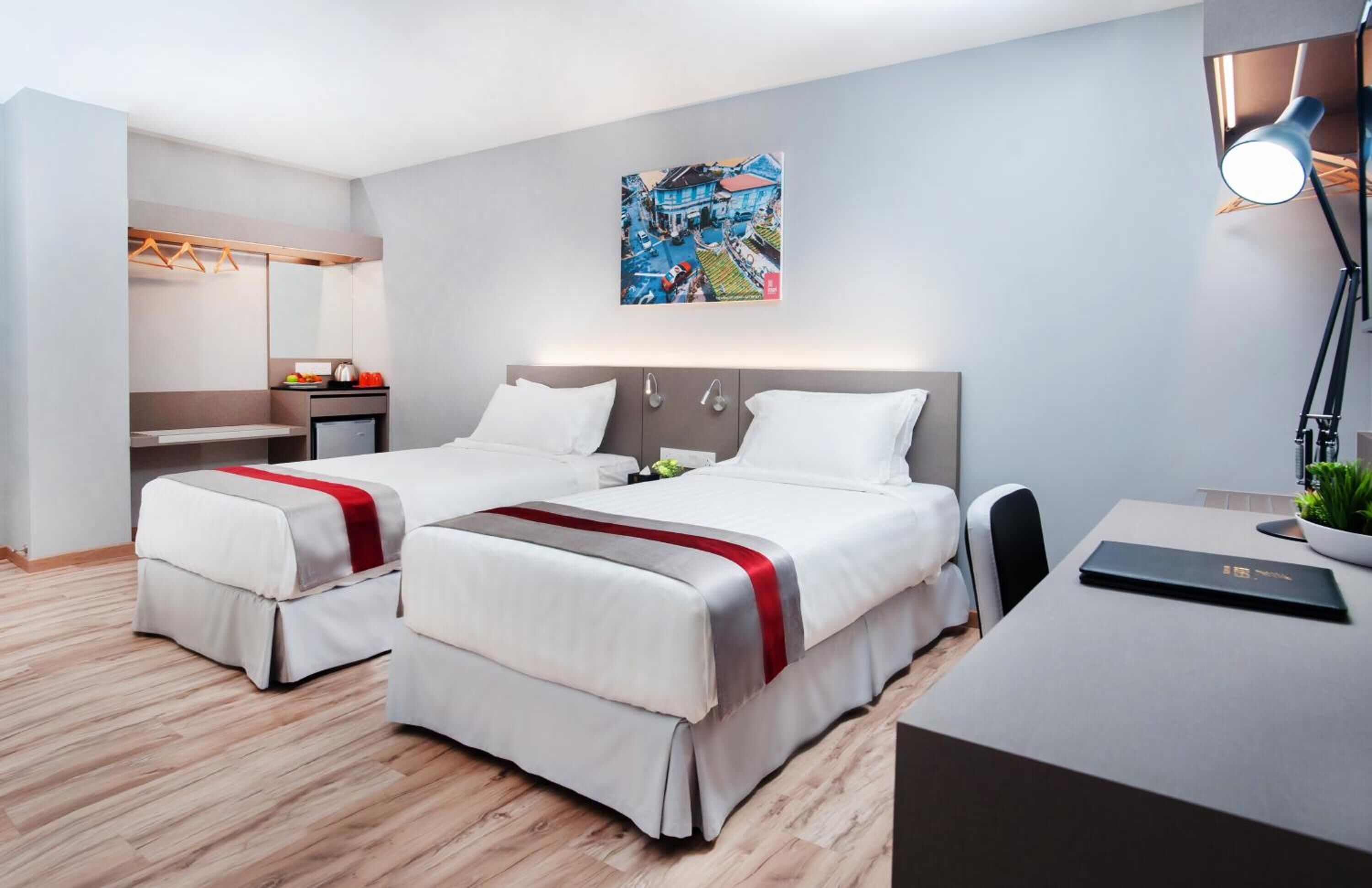 A modern hotel room with two beds, a desk, a chair, a small table with a plant, a mirror, and a painting on the wall.