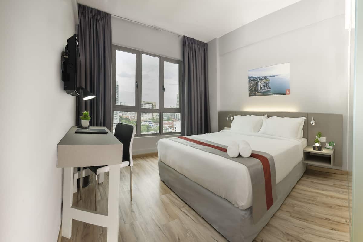 A modern hotel room with a large bed, a desk with a chair, a TV mounted on the wall, and a city view through the windows.