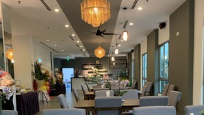 Ein modernes Restaurant mit Holztischen und grau gepolsterten Stühlen, einer zentralen Chandelüster und einer Bar im Hintergrund.