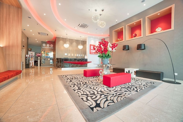 Un hall de hotel con mostrador de recepción, asientos rojos, alfombra estampada y estanterías decorativas.