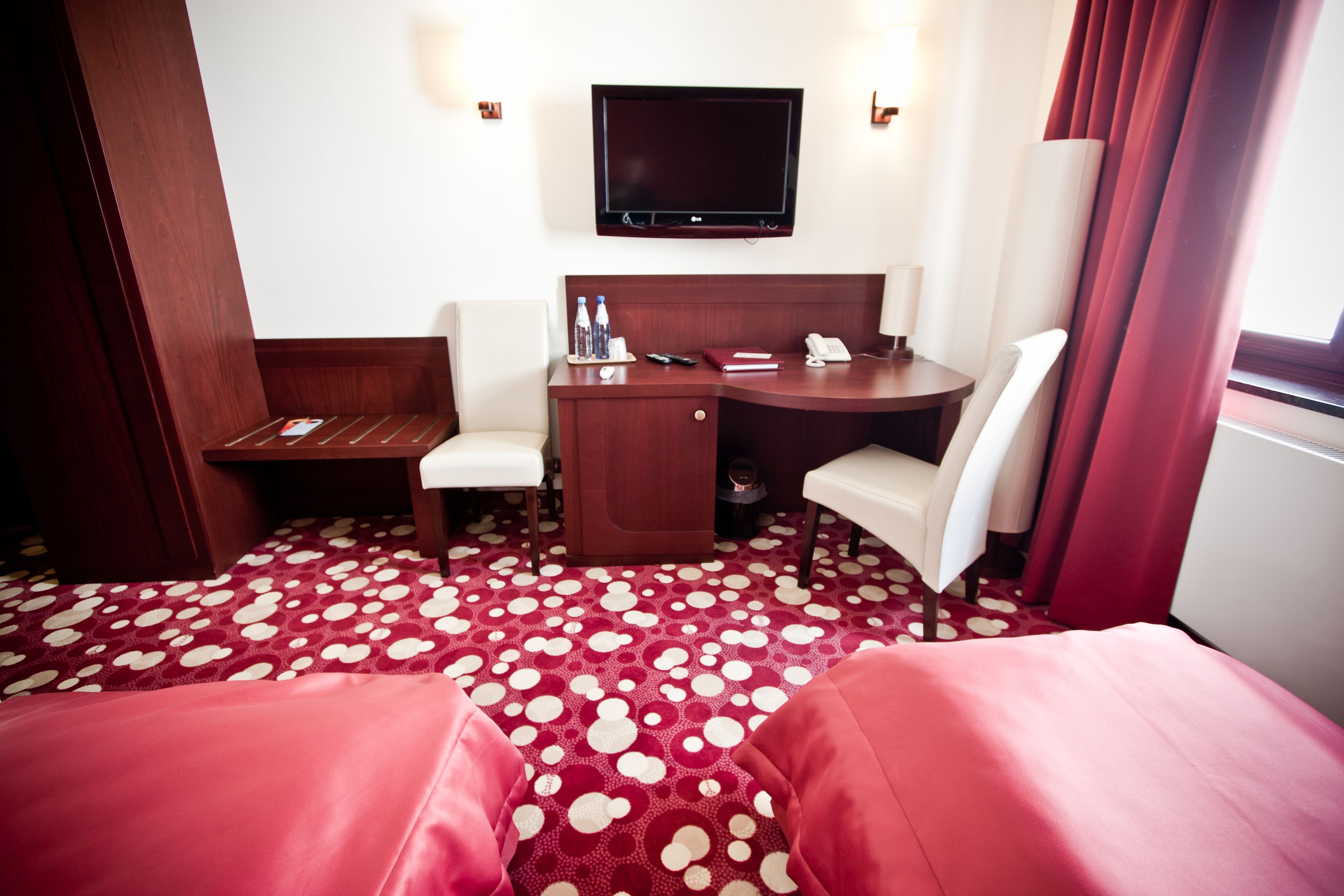 Ett hotellrum med en säng, ett skrivbord, en stol, en tv och en röd matta med prickar.