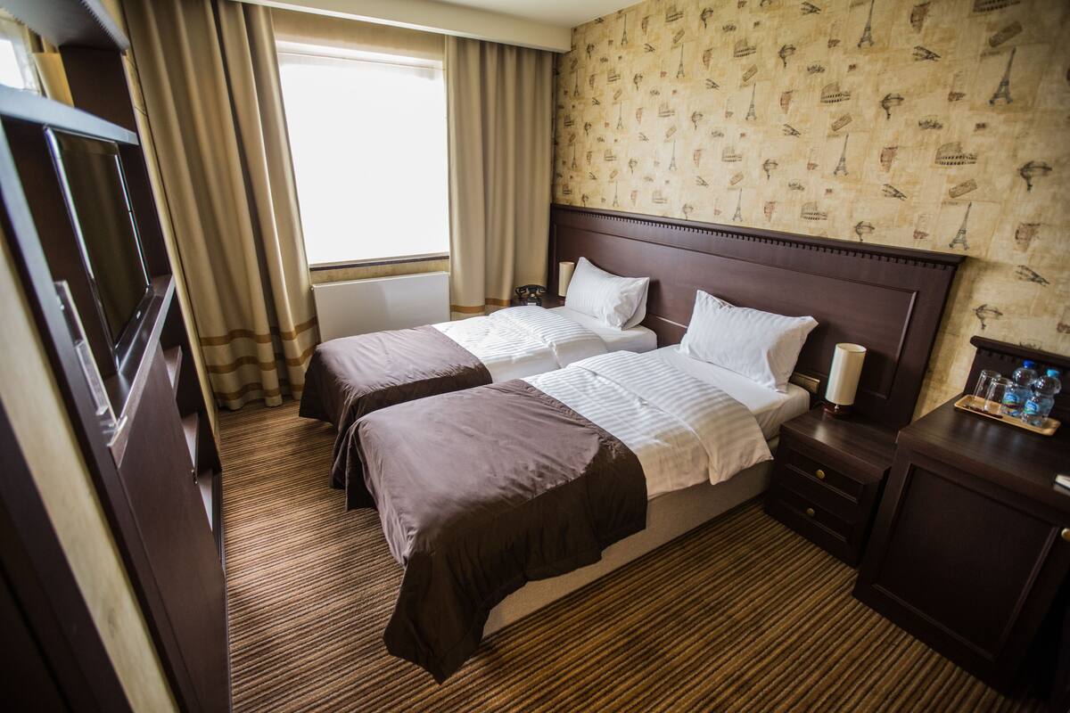 A hotel room with two beds, a TV, a nightstand, and a window with curtains.