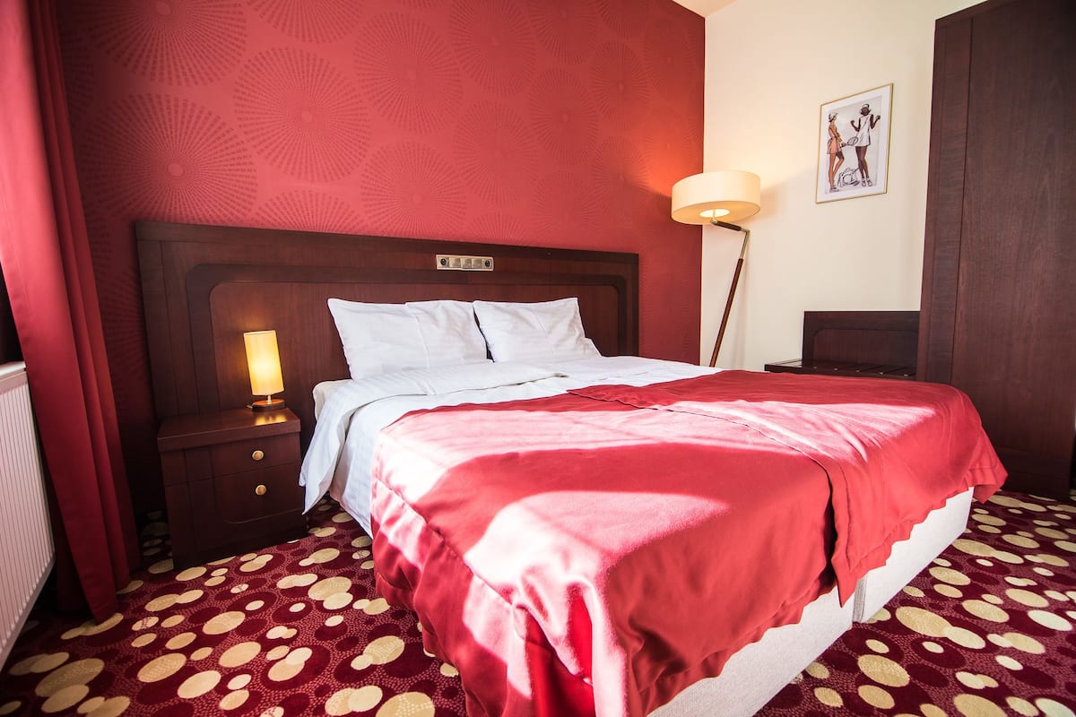 A hotel room with a large bed, a bedside table with a lamp, a red carpet, and a wall with a circular pattern.