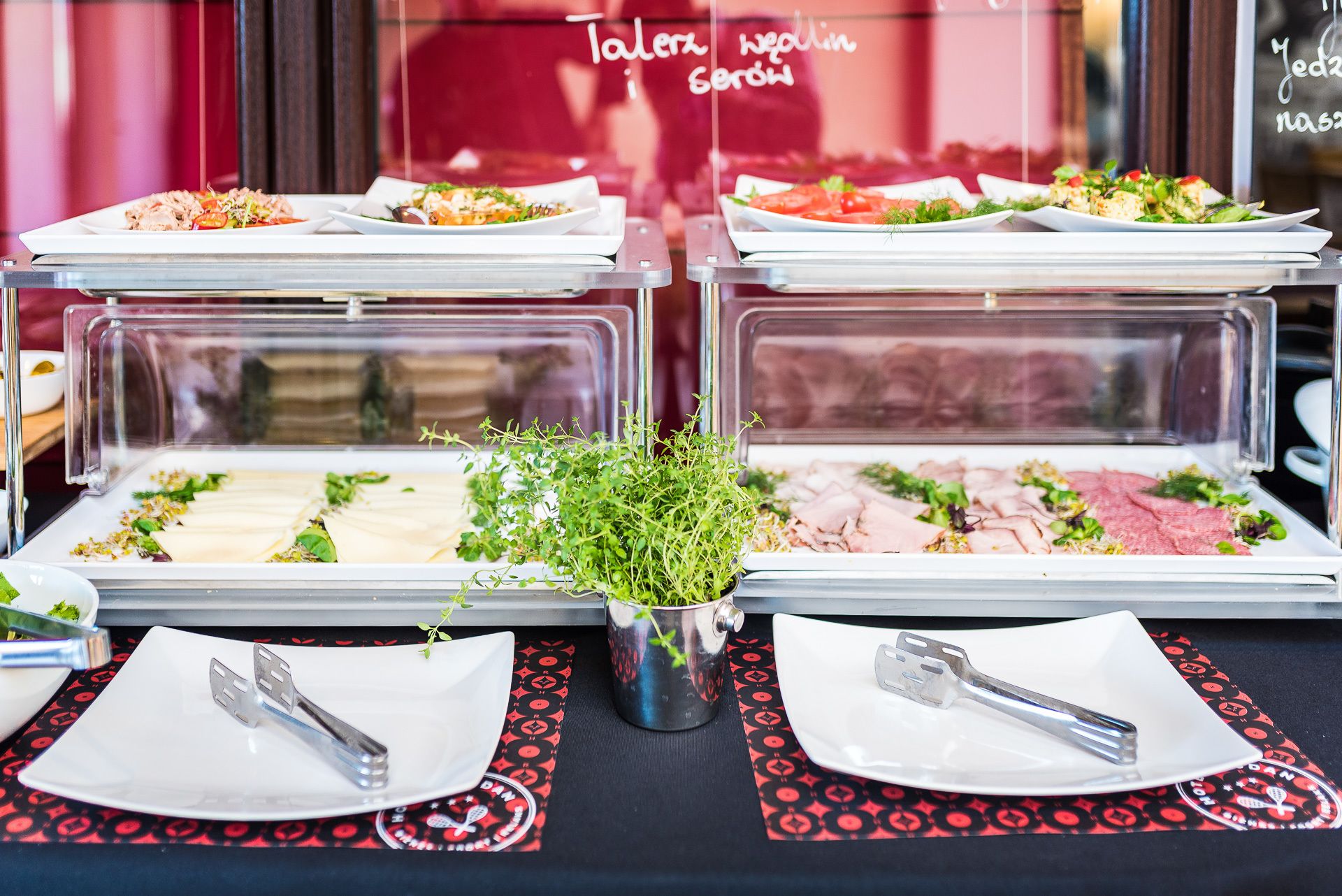 Montaje de bufé con diversos platos, entre ellos ensaladas, carnes y guarniciones, presentados en recipientes transparentes.