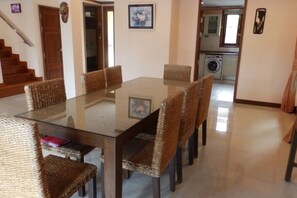Een eethoek met een tafel met glazen blad, omringd door rieten stoelen, een trap en een keuken op de achtergrond.