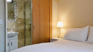 A hotel room with a bed, a nightstand with a lamp, a wooden wardrobe, and a glass shower enclosure. - White House Cottages (Durham)