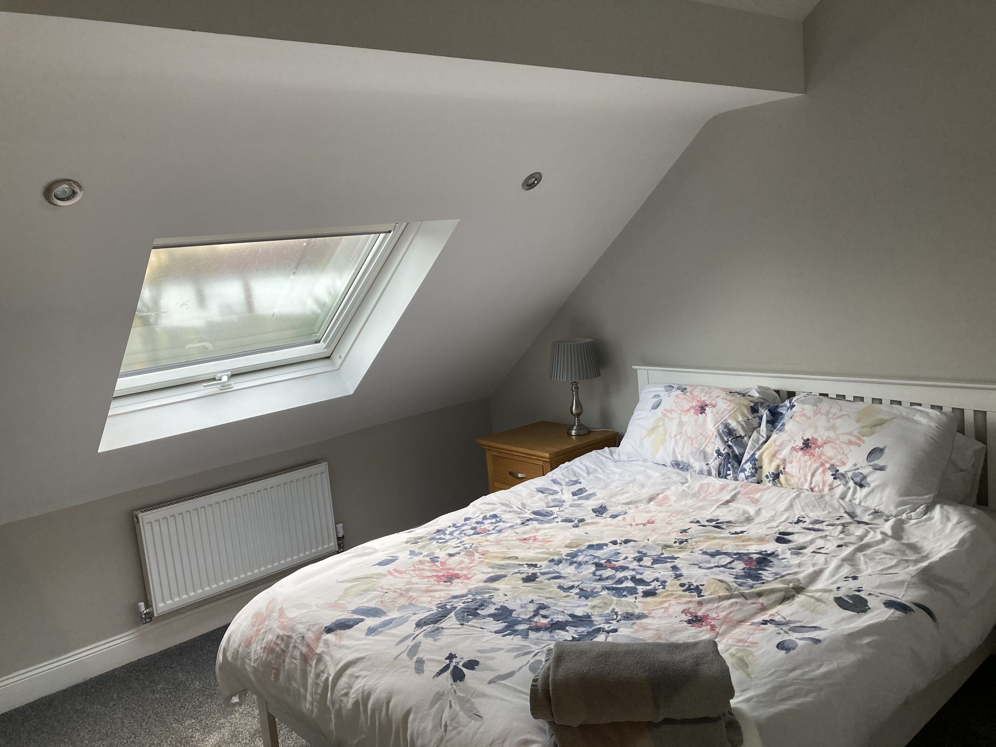 Ein Schlafzimmer mit Dachfenster, einem Bett mit floralem Bettzeug, einem Nachttisch mit Lampe und einer Heizung.