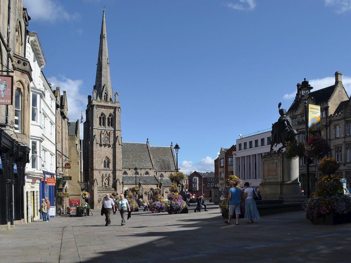 A historic town square with a prominent church tower, a statue, and flower arrangements.
