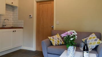 A compact living space with a kitchenette, a grey sofa with patterned cushions, a small table with a vase of flowers and a bottle of wine, and a door leading to another room.