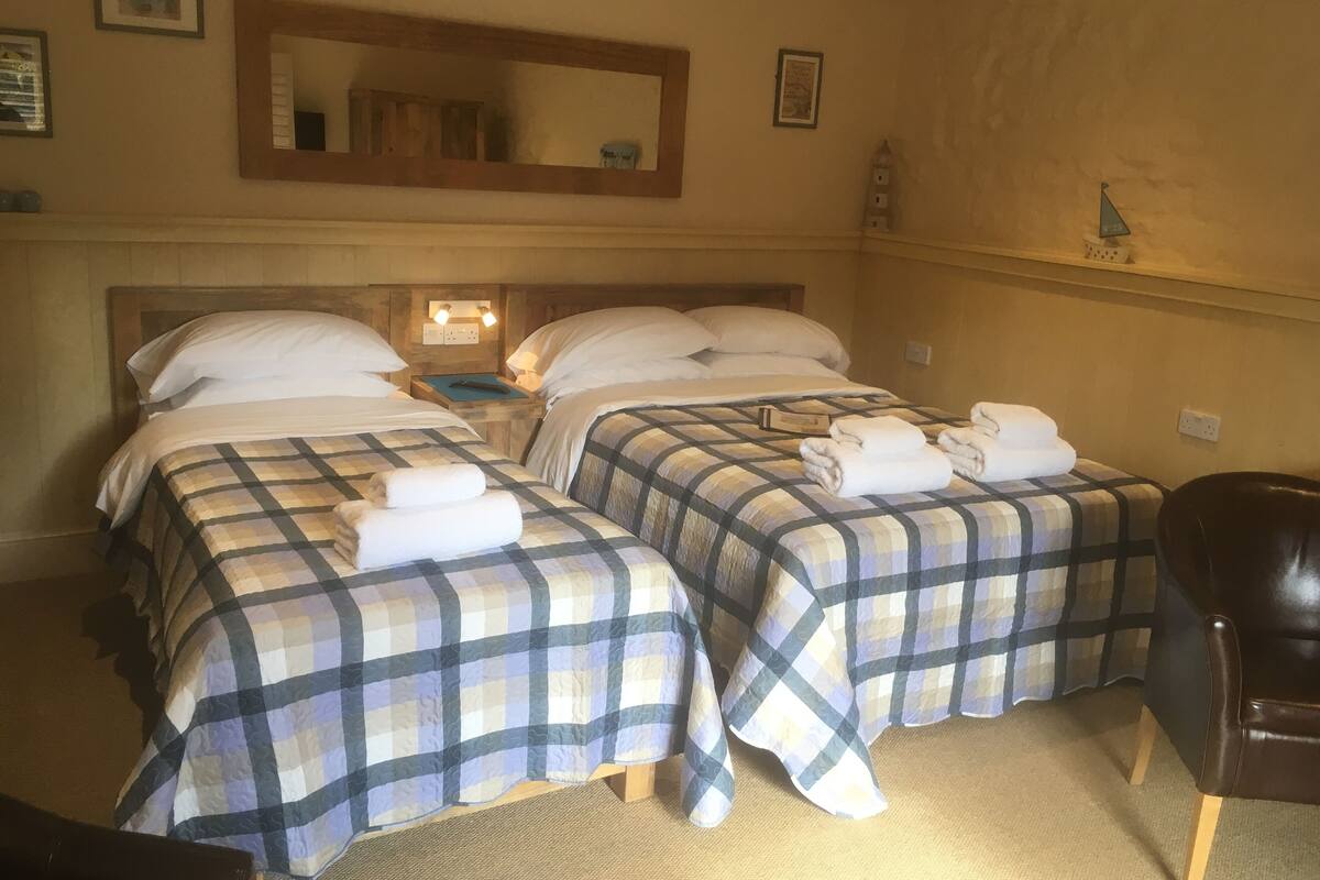 Hotelkamer met twee bedden, elk met een geruite sprei en kussens, een nachtkastje met een lamp, een stoel en een klein plankje met decoratieve voorwerpen.