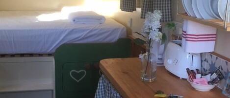 A small, cozy room with a bed, a wooden table with kitchen items, and a shelf with plates.