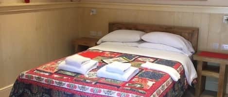 Una cama bien hecha con una colcha estampada y toallas dobladas, una mesita de noche de madera con una bandeja roja, y un espejo en la pared.